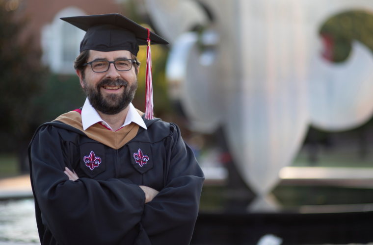 A man is pictured wearing a black cap and gown near a fleur de lis sculpture in UL Lafayette's quad.